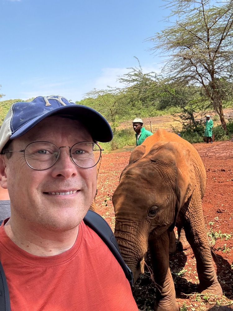 A white man wearing glasses and a baseball cap takes a selfie with a young elephant in the background. An elephant is standing very close behind him, with caretakers wearing green in the background.
