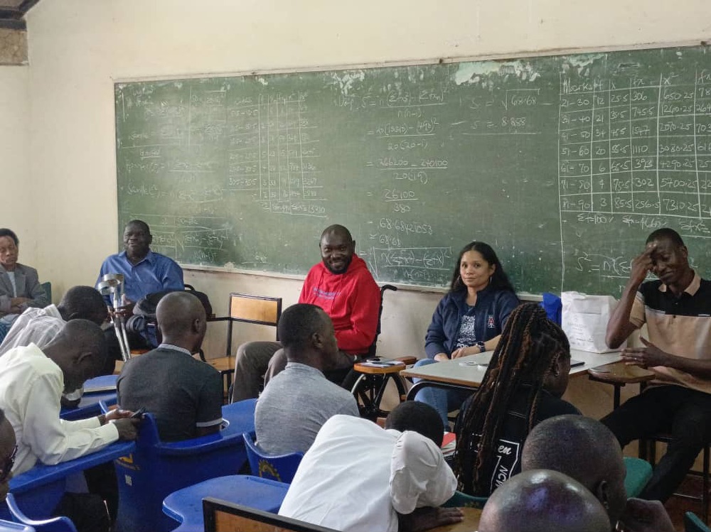 A group of people, mostly Ugandan men and one American woman, seated in a classroom. There is a blackboard in the background.