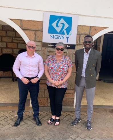 A white American woman stands between two Kenyan men in front of a building with a sign that reads "Signs TV" behind them.