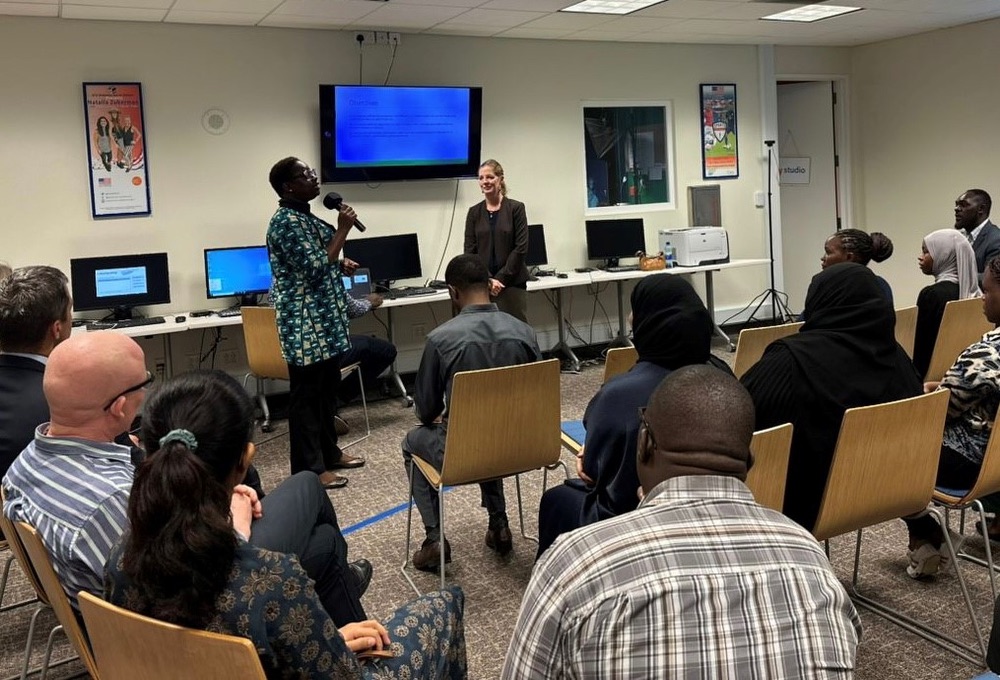 Two women, one American woman (back right) and one Tanzanian woman (back left), presenting to an audience in front of a conference room. A TV screen is visible in the back, with a row of computer desks along the wall. People are seated on chairs in the audience. 