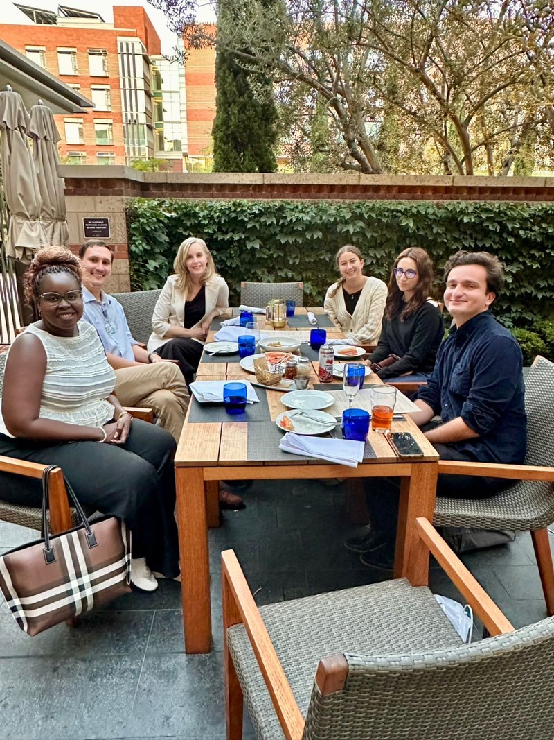 Six people sit outdoors at a table having lunch, smiling at the camera.