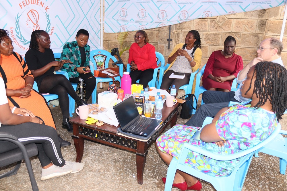 A group of people, predominantly women, are seated in a circle outdoors, engaged in discussion. The background features a banner with the logo and name of “FedWen-K” (Federation of Deaf Women Empowerment Network).