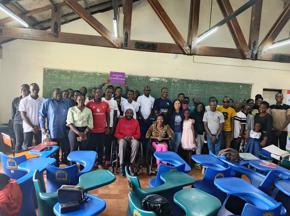 A group of people, mostly Ugandan women and men, one American woman, and a few children, posing for a group photo in a classroom.