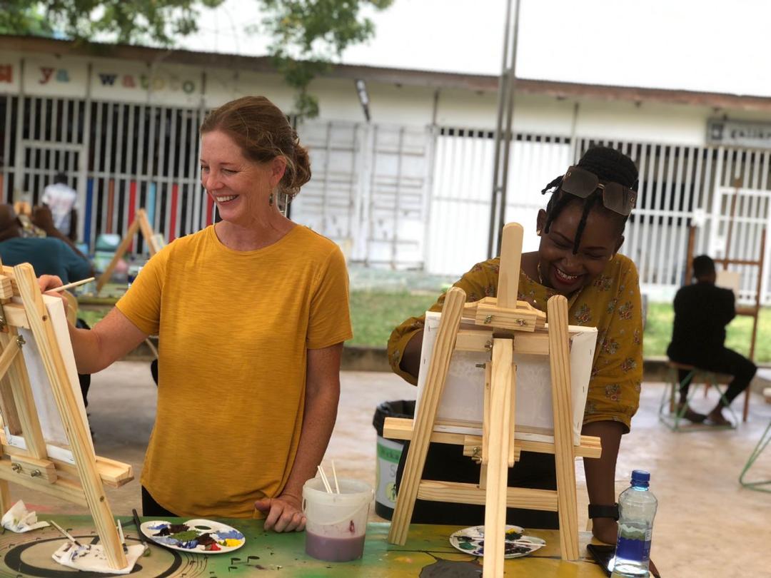 A white, American woman and an Tanzanian woman standing next to one another, each painting on an easel and smiling. 