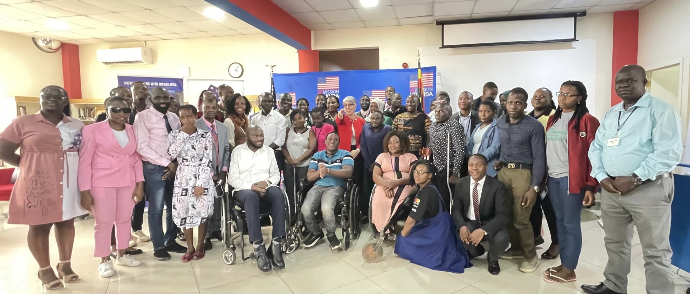 A large group of men and women from Africa, some who are in wheelchairs and others who are standing, pose for a picture in front of a banner that reads US Mission Uganda. 