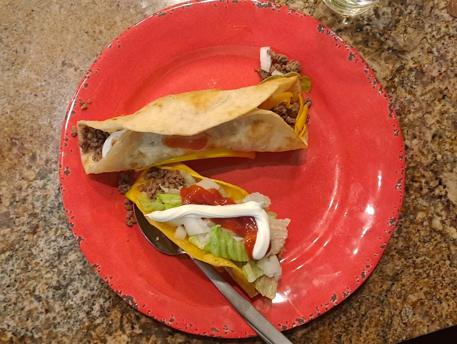 Two tacos on a red plate with ground beef, lettuce, cheese, sour cream and salsa. One Taco is folded, and the other is open with a spoon placed underneath.  