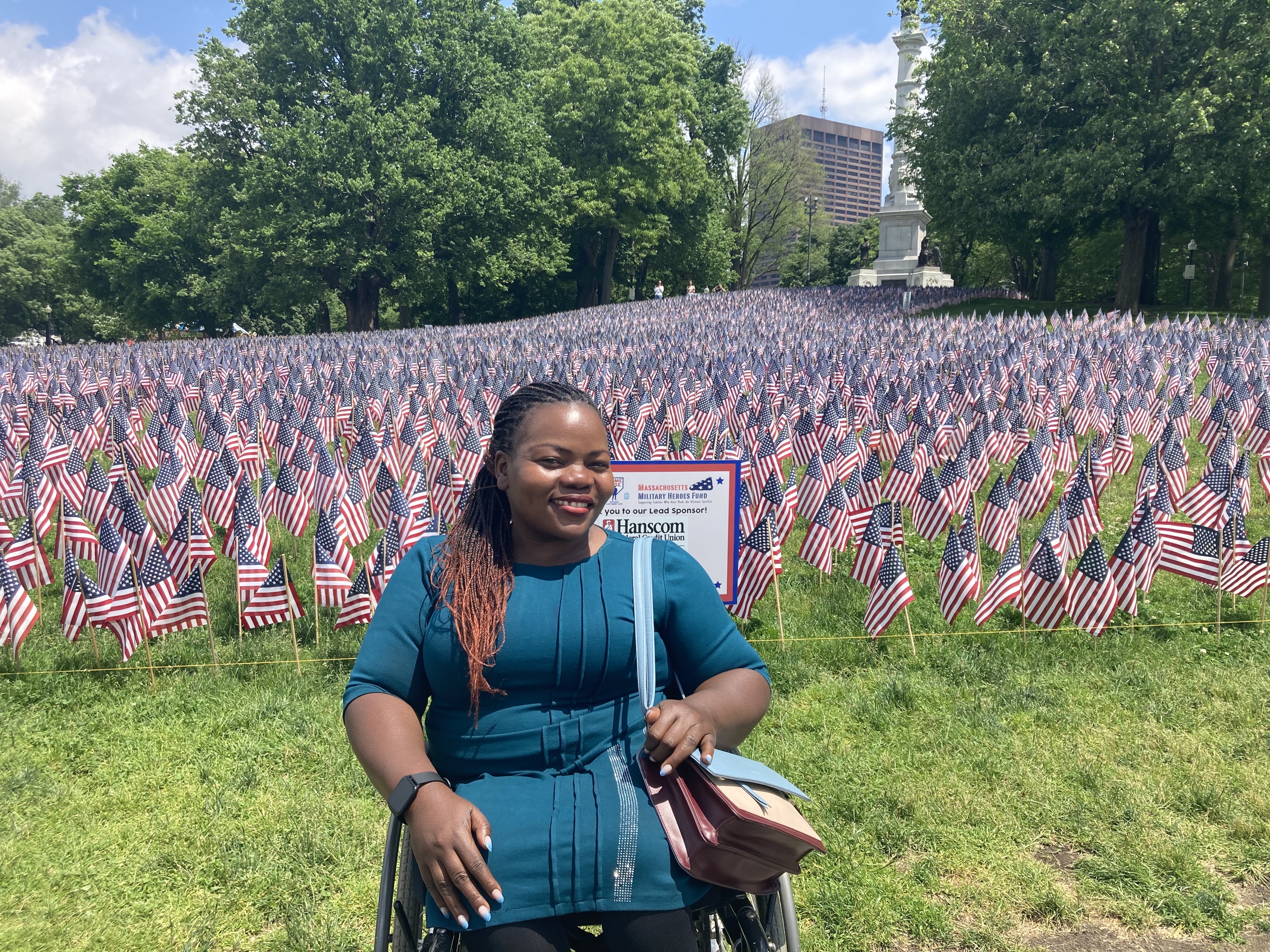 A Ugandan woman with long braided hair, wearing a blue dress, holding a handbag, poses in a wheelchair in front of American flags.