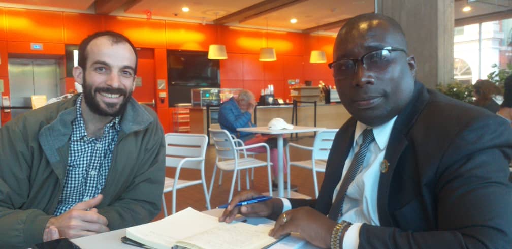  Two men are sitting at a table, likely in a cafe, and smiling at the camera. The man on the left is American with short hair, a full beard, wearing a striped shirt and a light grey jacket. The man on the right is Tanzanian with short hair, wearing glasses, a white shirt with a tie, and a black suit. He is also holding a pen. 