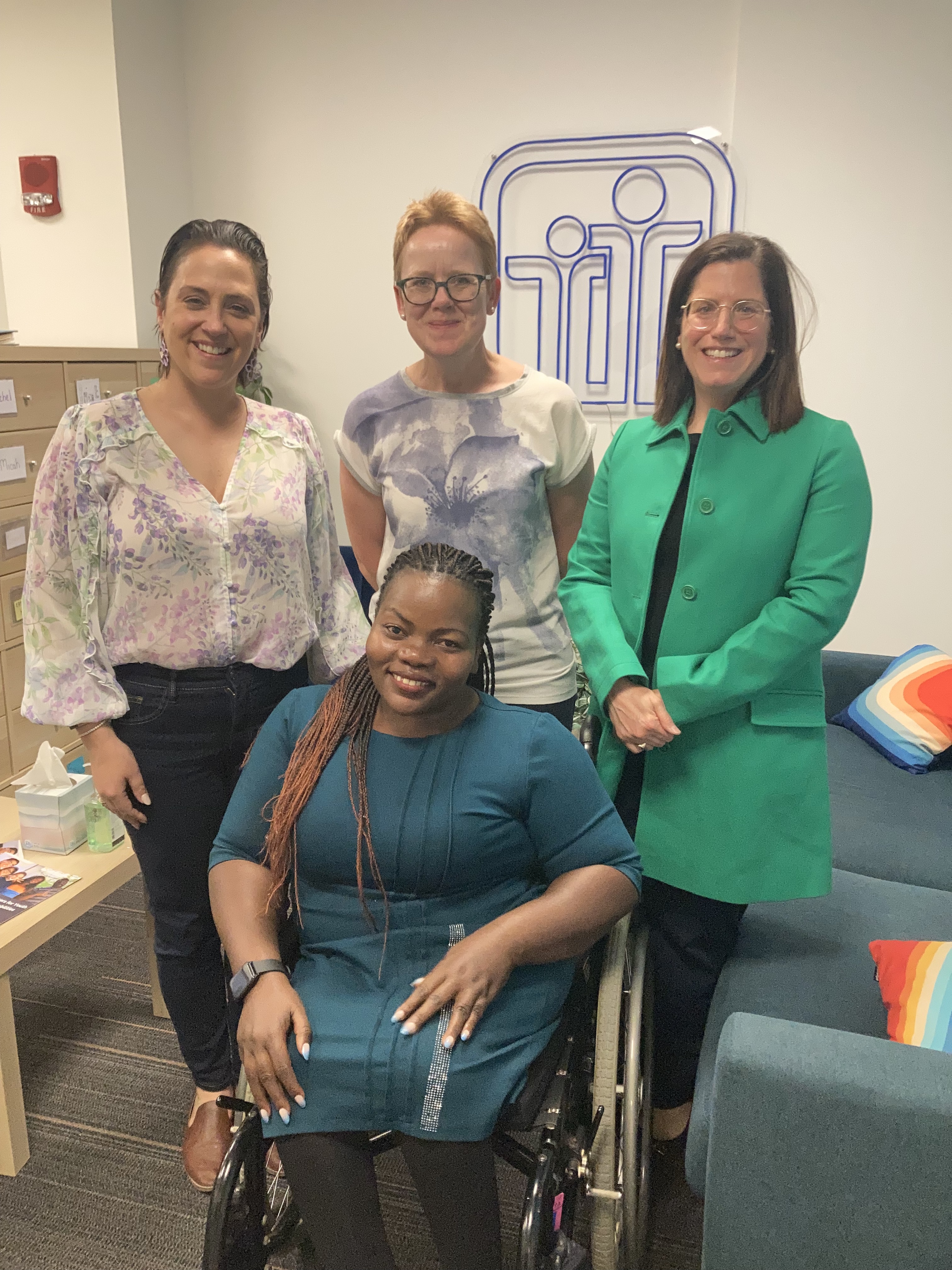 Four women, three American and one Ugandan, pose for a photo, all smiling. The woman on the left wears a patterned blouse and black pants, the one in the middle wears a white blouse and black pants, and the woman on the right wears a green jacket and black pants. The Ugandan woman in front of them, sitting in a wheelchair, wears a blue dress.