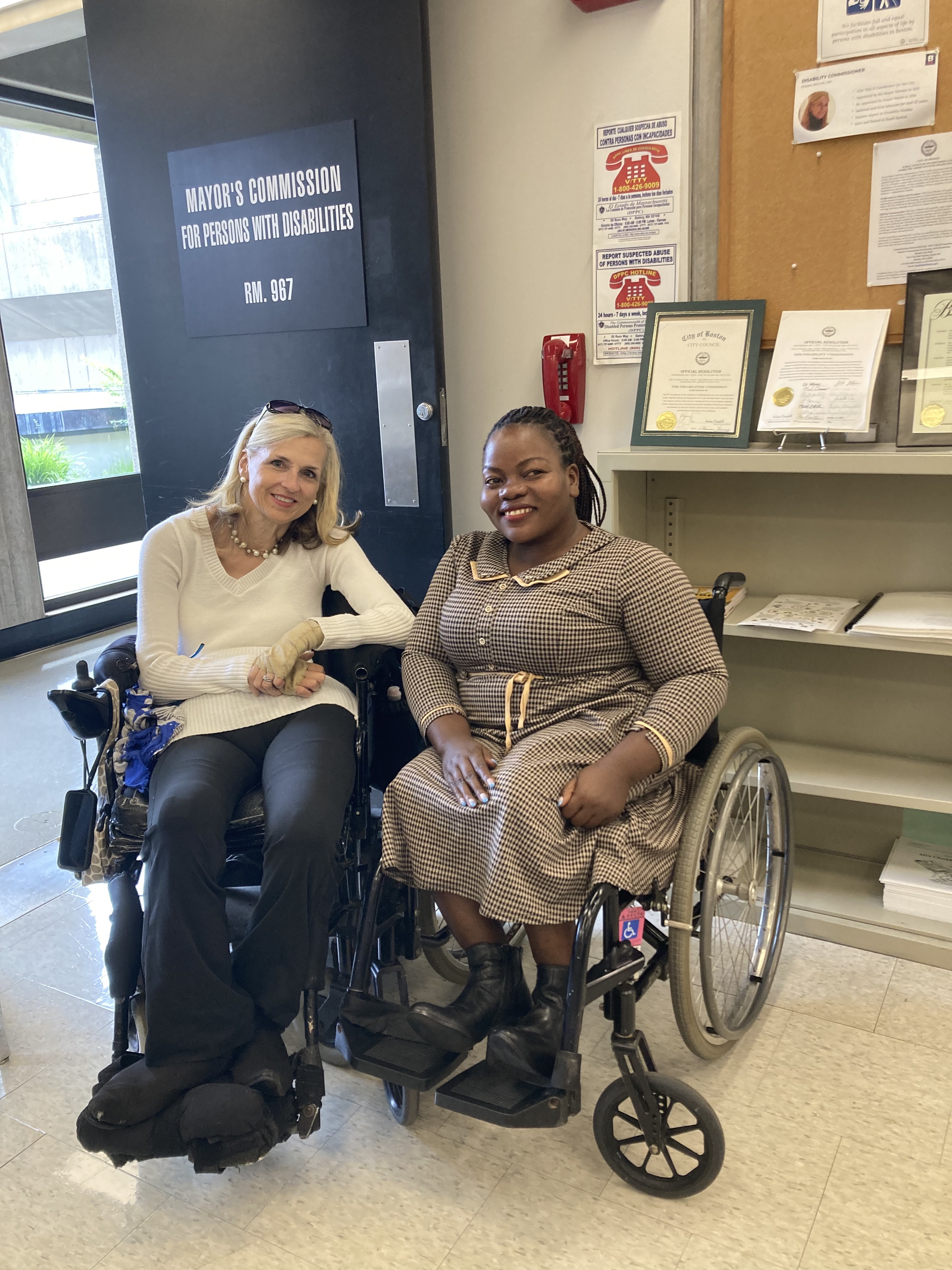 Two women in wheelchairs, one American and one Ugandan, pose for a photo beside a door labeled "Mayor’s Commission for Persons with Disabilities" Room 967, both smiling. Behind them is a shelf and a notice board with certificates. The woman on the left is wearing a white top and black pants, and the woman on the right is wearing a brown long-sleeved dress.