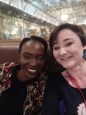 An African woman wearing a patterned jacket and black shirt poses with a white woman with short black hair wearing a navy top. 