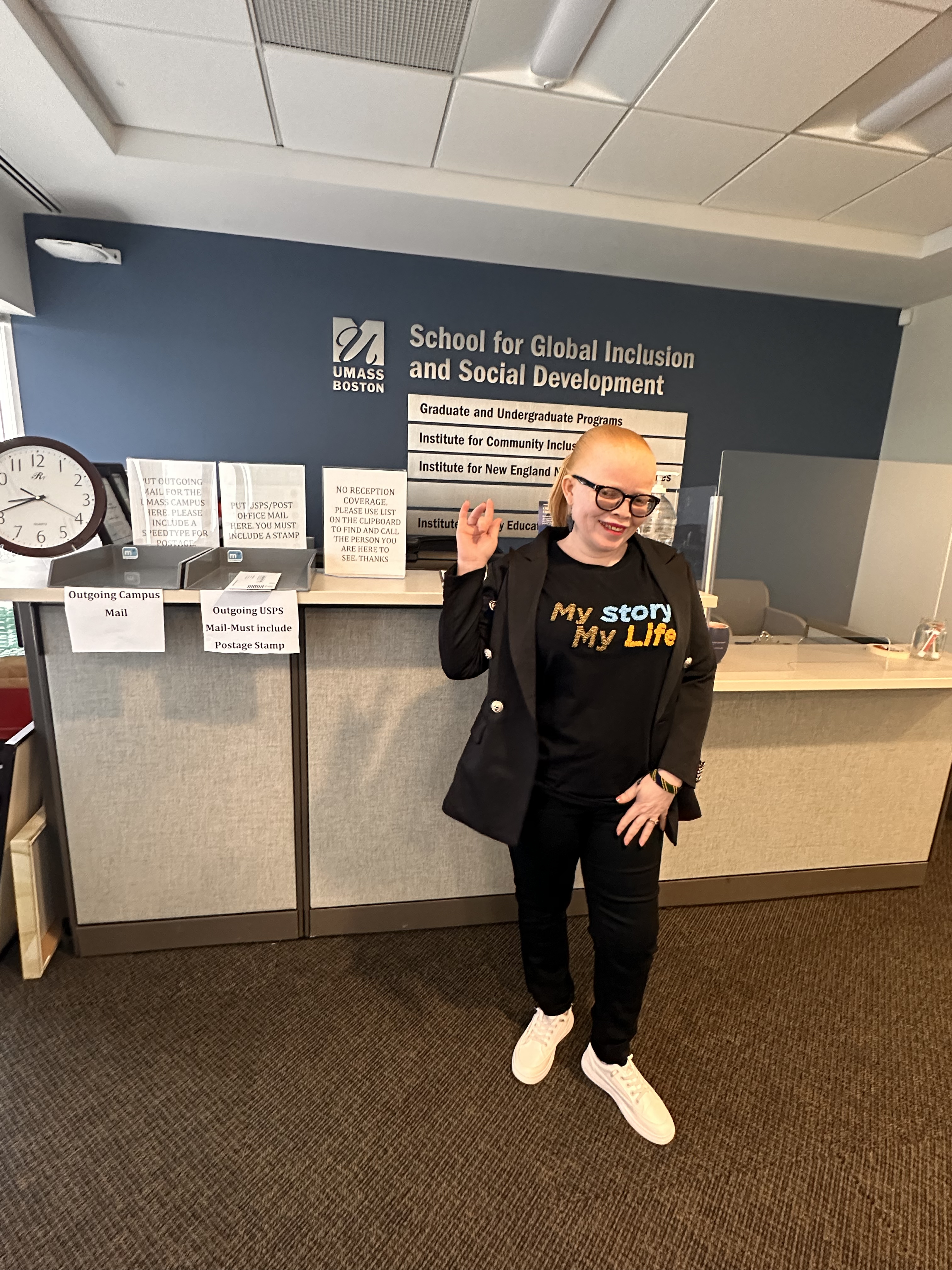 Alt Text: A light skinned woman with red hair, wearing black glasses, black pants, and a black shirt that reads “My Story, My Life,” stands in front of an office desk. 