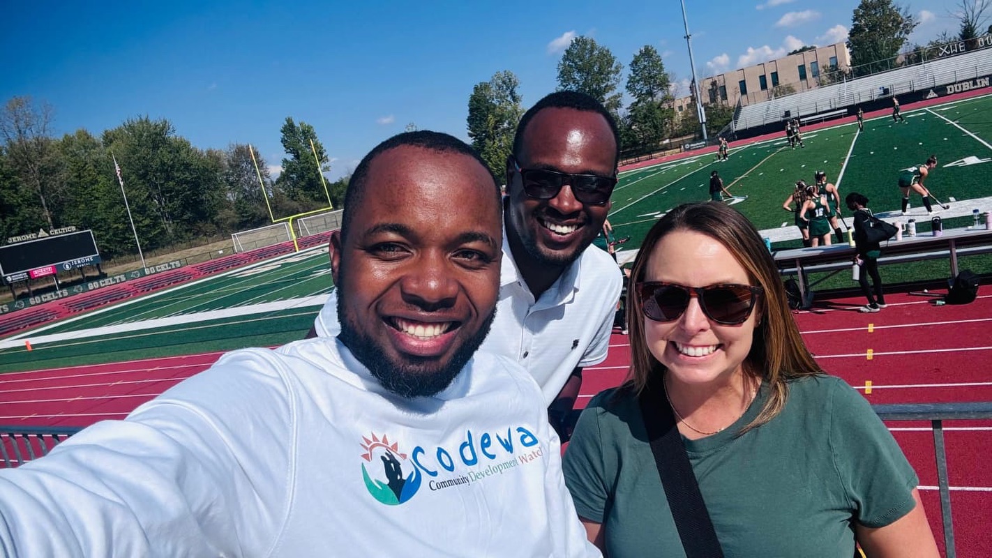 Two men and a woman stand together on a sports field, smiling as they take a selfie.