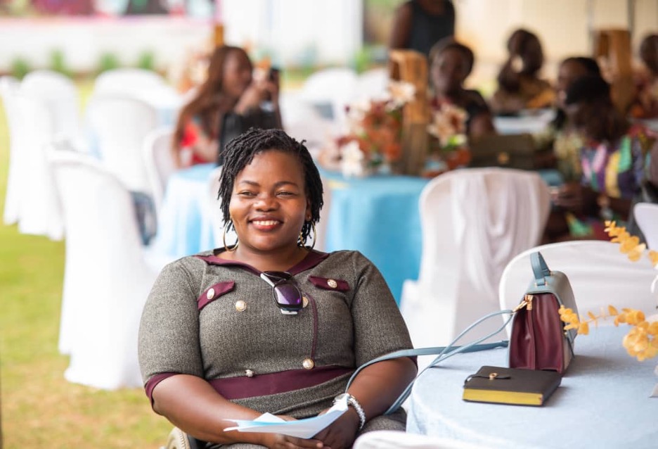 A Ugandan woman with braided hair, sitting at a white cloth-covered table in an outdoor setting, smiling.