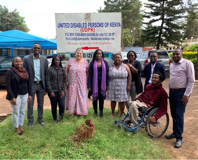 A white American woman stands outdoors amidst a group of Kenyan women and men, including one man using a wheelchair. They are gathered in a partially grassy and paved area. In the background is a sign with the words “United Disabled Persons of Kenya (UDPK)” along with the organization's address and slogan, which is “the voice of persons with disabilities.”