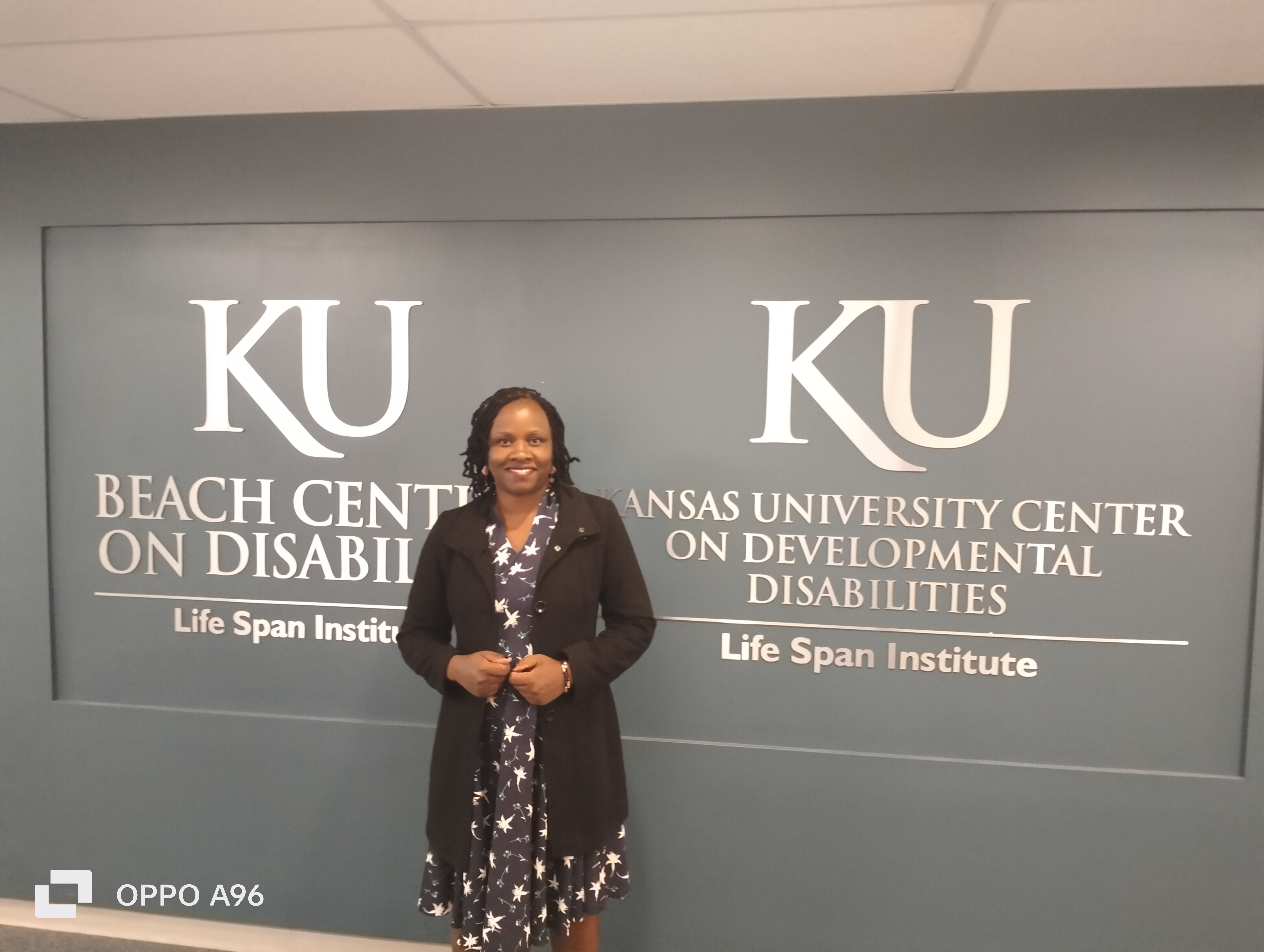 Alt-Text: An African woman with long black hair, wearing a long black coat, stands in front of a wall with the words "KU Beach Center on Disability" and "KU Kansas University Center on Developmental Disabilities."