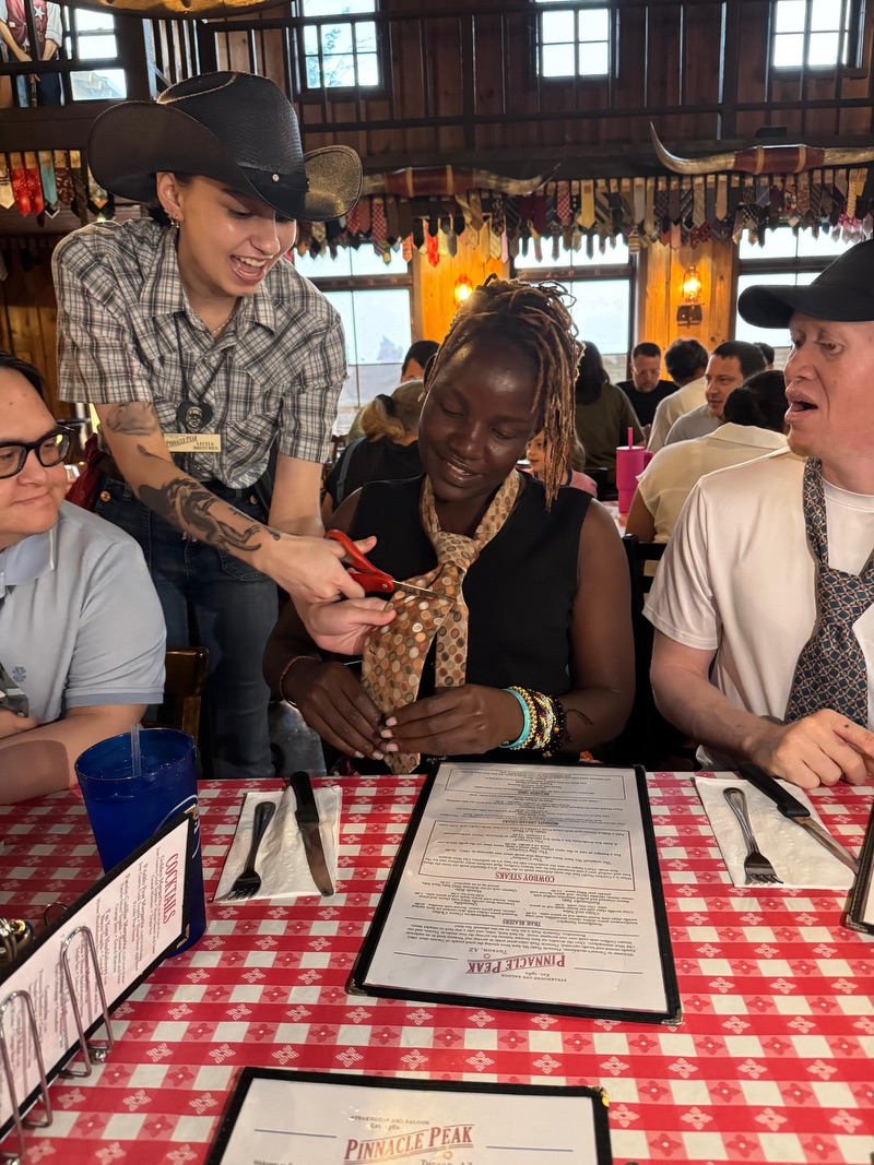 A woman seated at a table with two men, smiling, while a server is playfully cutting her tie as part of the celebration.