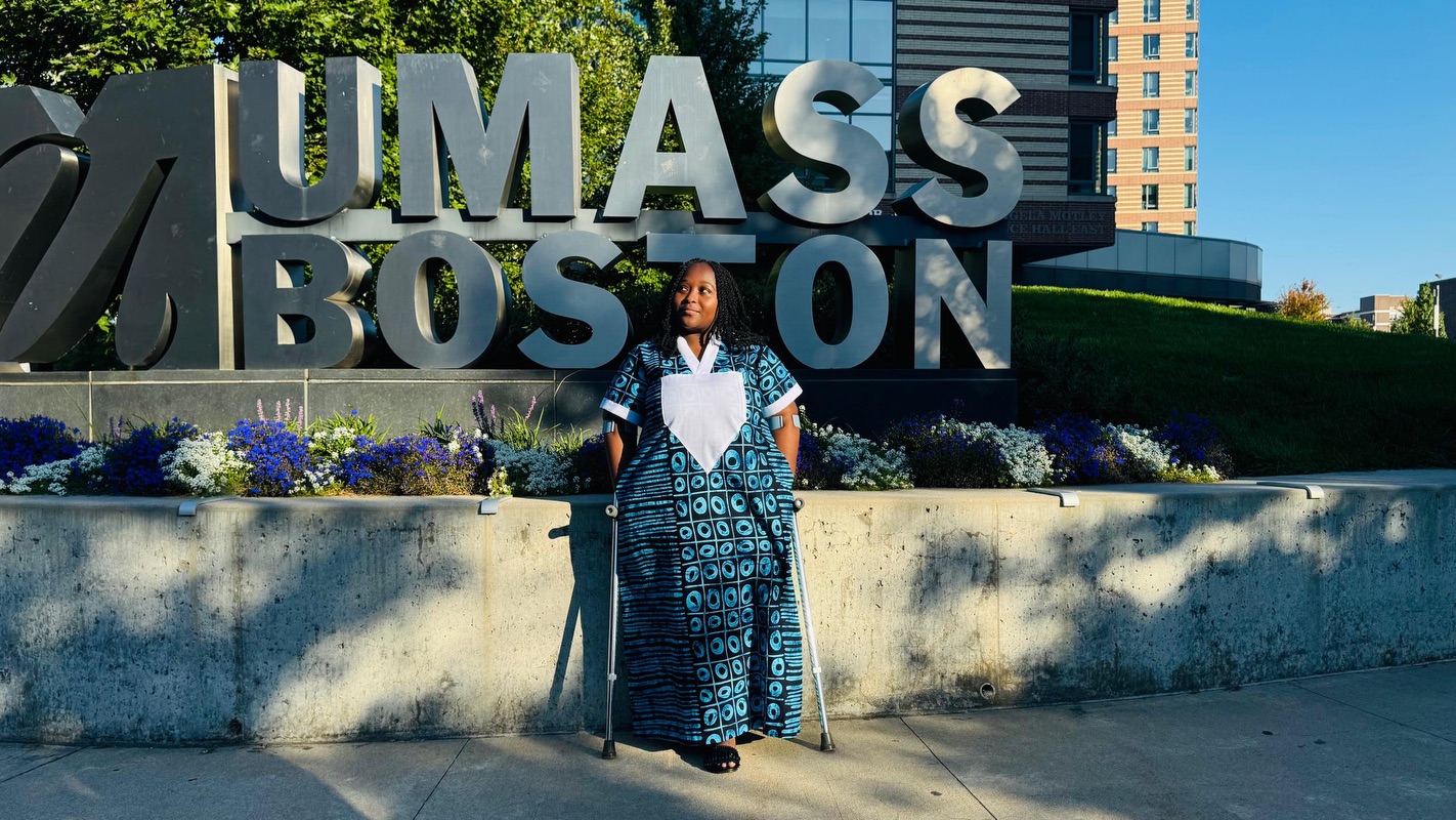 A young woman with a colorful dress and crutches stands in front of a sign that says “UMass Boston.”