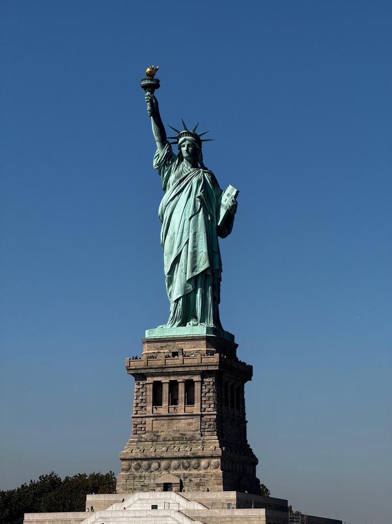 A green statue of a robed woman representing the Roman goddess of Libertas. She holds a torch in her right hand, raised up above her head. She holds a tablet in her left hand. She wears a seven-pointed crown.