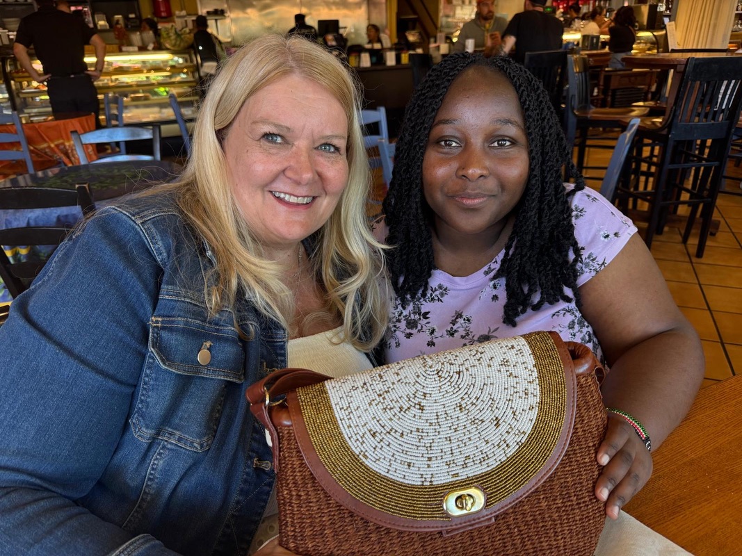 Two women seated at a table, smiling at the camera; one is holding a handmade handbag.
