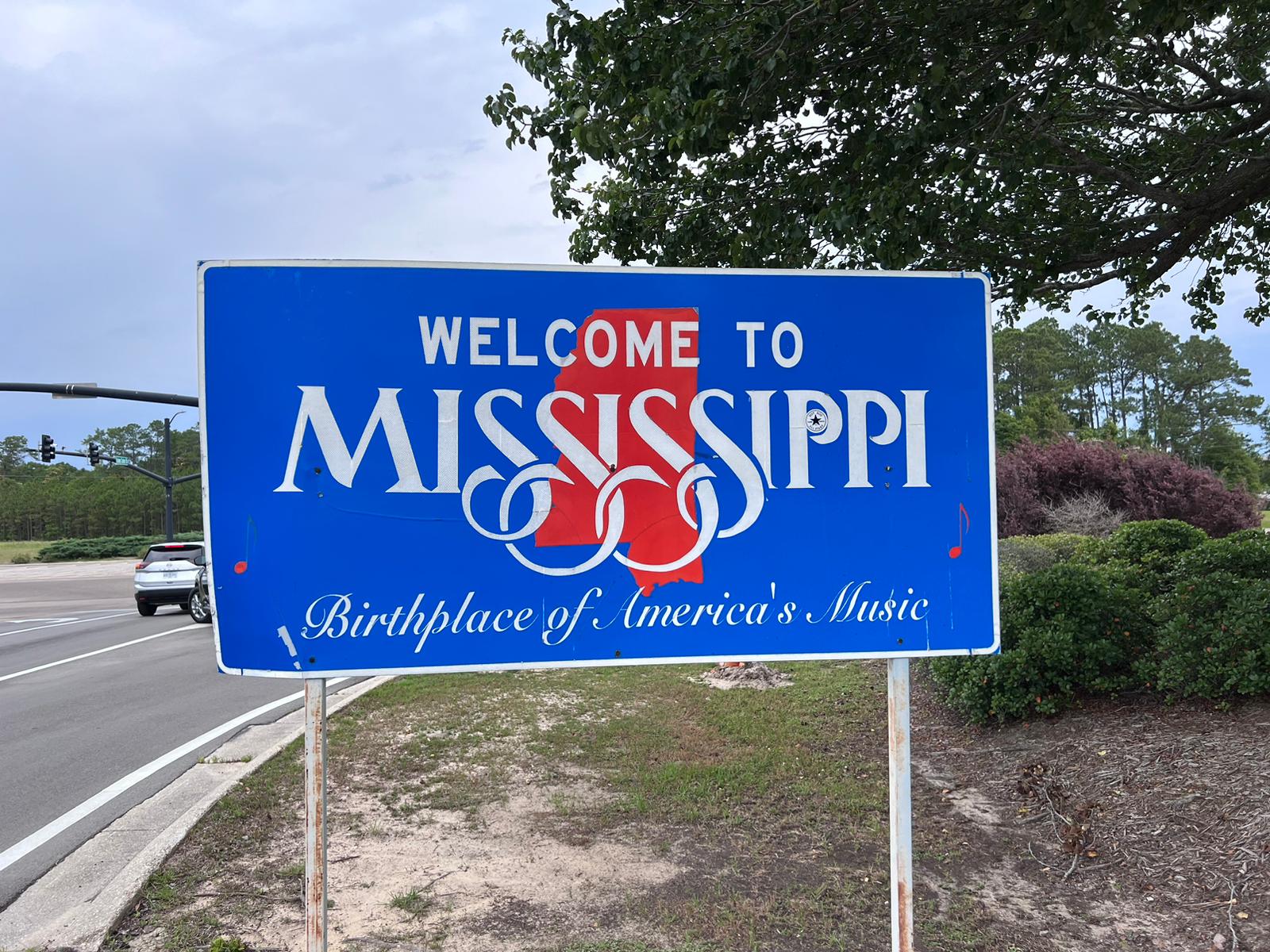  A blue road signage with white texts that read “Welcome to Mississippi” and “Birthplace of America’s Music” The sign is positioned near a road with trees and shrubs in the background. 