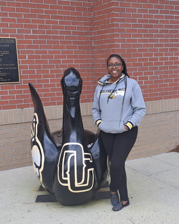 An African woman wearing a gray jacket with black and gold accents and black pants stands beside a black statue of a bird with two logos painted on its body. The statue is positioned outdoors in front of a brick wall.