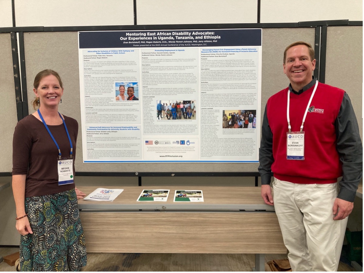 A white woman (left) and a white man (right) posing in front of a poster mounted on a poster board and a table with materials, both smiling into the camera. 