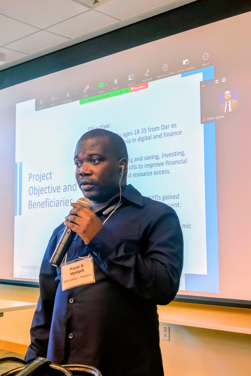 A man wearing earphones holds a microphone while presenting in front of a screen displaying a PowerPoint slide.