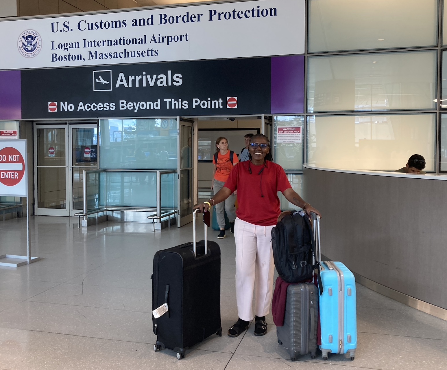 A woman with several suitcases walks out of the airport arrival gate, with other travelers behind her.