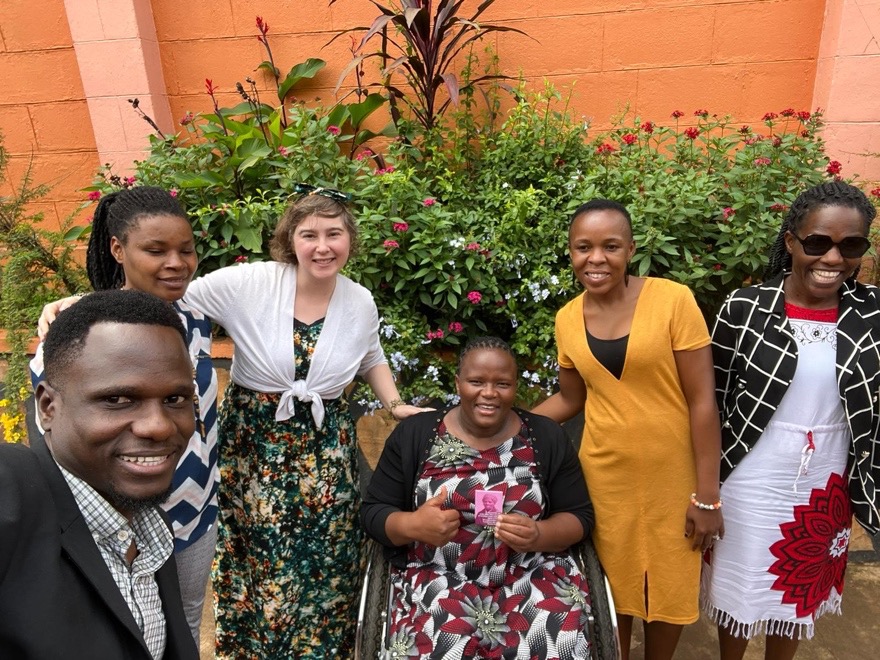 A group of six people, including one white American woman, four Ugandan women, and one Ugandan man, posing for a picture. One of the Ugandan women uses a wheelchair. 