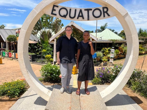 A white, American woman and a Ugandan woman standing inside a stone circle with a sign above them that reads “Equator,” both smiling into the camera.