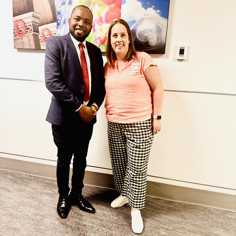 A man and a woman posing together for a photo, standing in front of a wall with artwork behind them. 