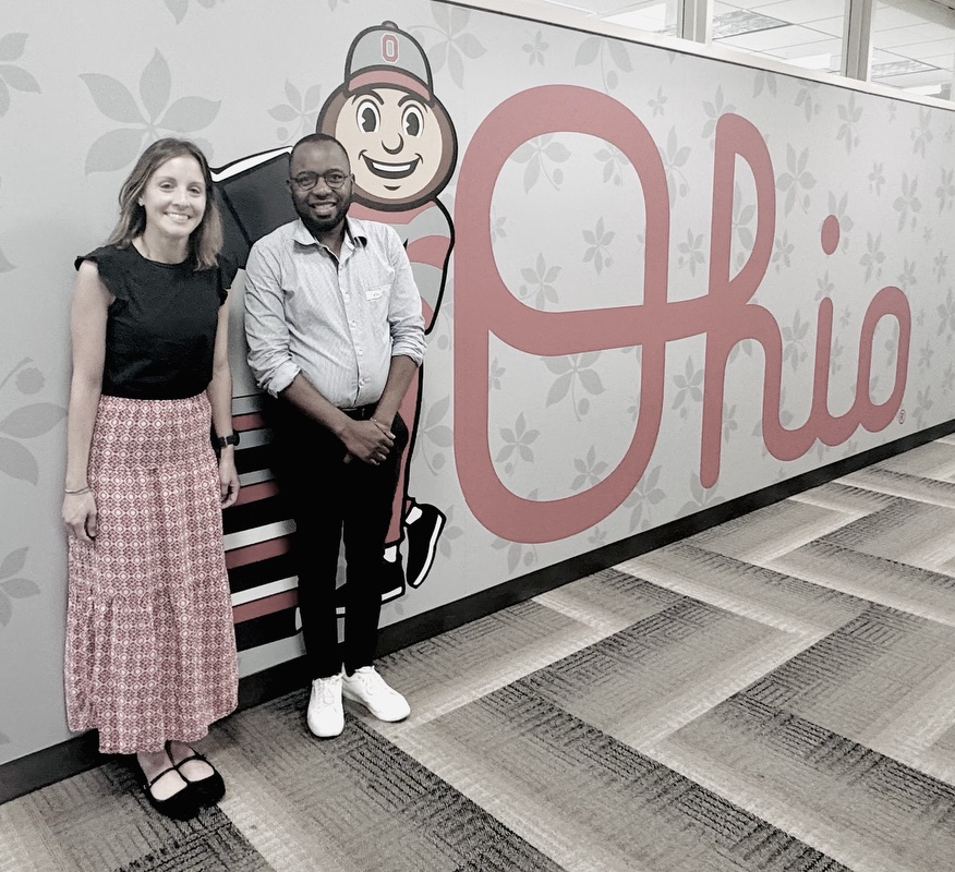 A man and a woman posing together for a photo in front of a wall decorated with a buckeye mascot and the word “Ohio.”