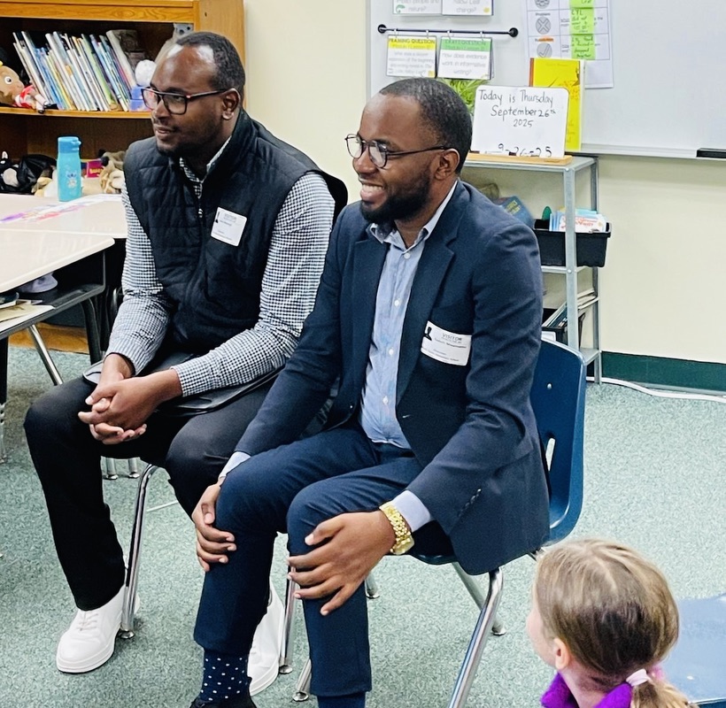 Two men seated on chairs in a classroom, with part of a student’s head visible in the corner of the photo.