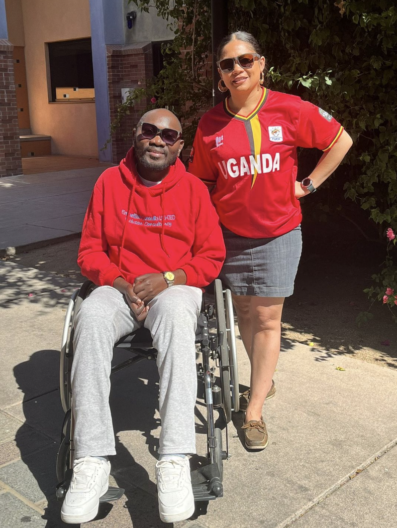 An African man in a wheelchair wearing sunglasses and a red sweatshirt stands beside a woman with tan skin, black hear and also wearing sunglasses and a red Uganda soccer jersey.