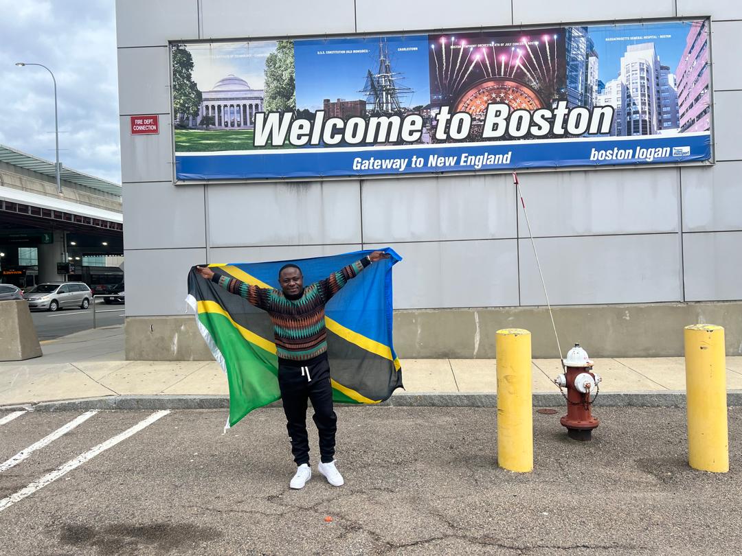 A Tanzanian man posing for the camera, holding the Tanzanian flag outside Boston Logan Airport after arrival. A “Welcome to Boston” billboard and a fire hydrant are visible in the background. 