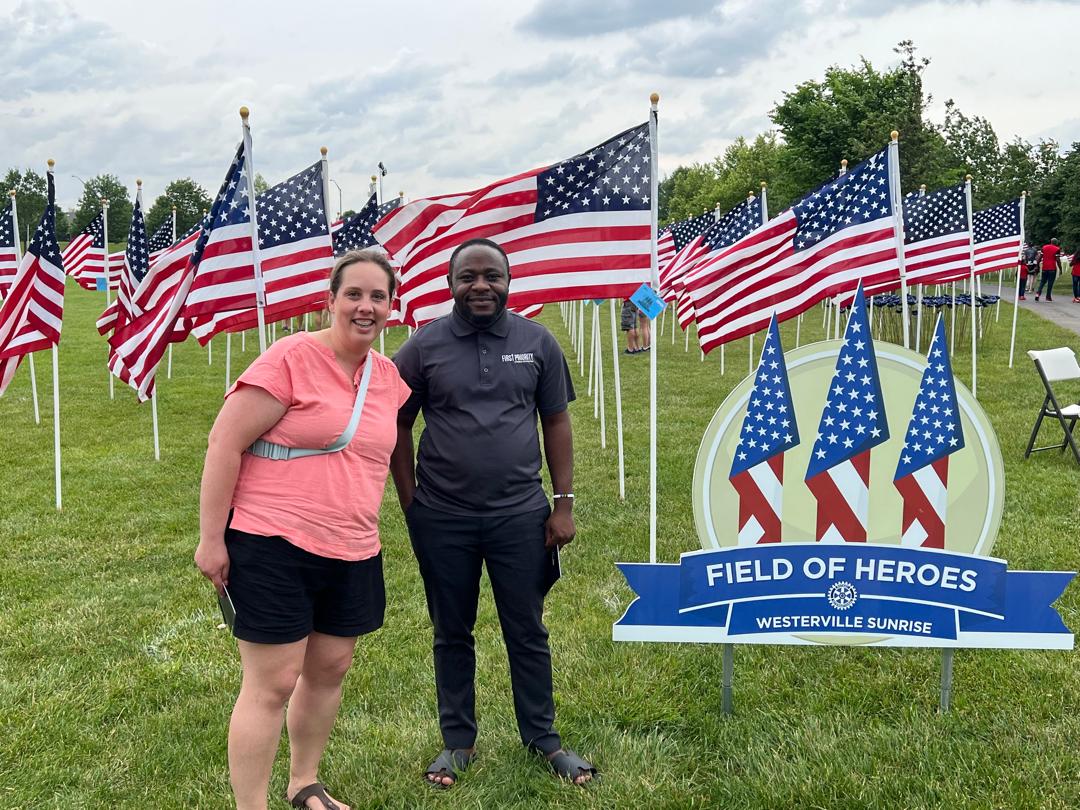 Two people, an American woman and a Tanzanian man, posing for a photo with many American flags behind them. There is a sign that reads “Fields of Heroes” next to them. 