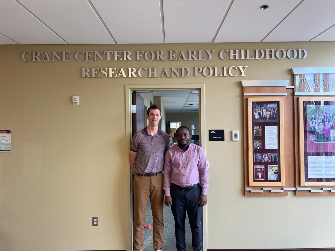 Two people, an American man and a Tanzanian man, posing for a photo outside an office. The words “Crane Center for Early Childhood Research and Policy” are visible on the wall behind them. Next to them is a glass case mounted on the wall displaying pictures and photos. 