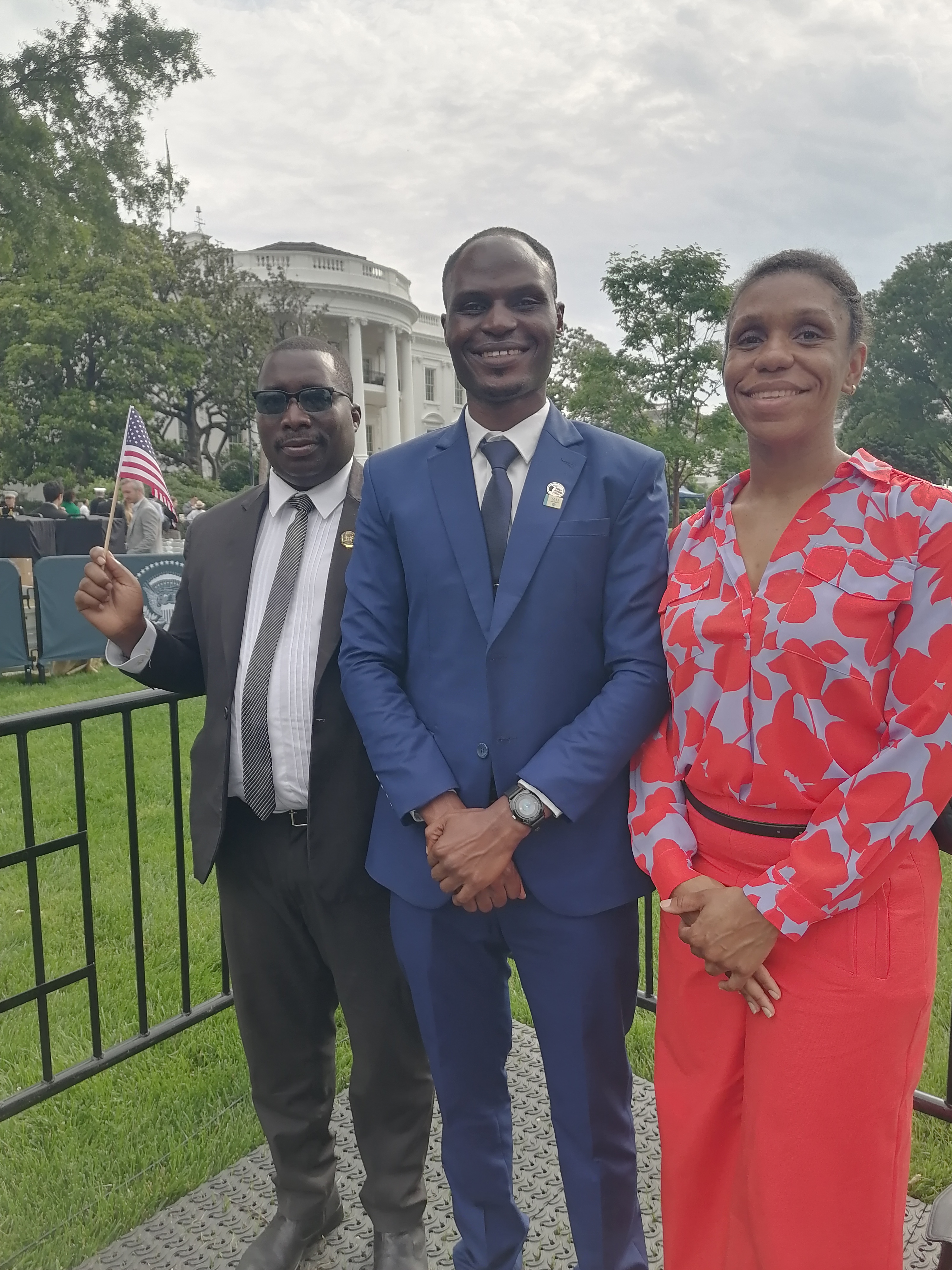 Alt text: From left to right, an African man wearing a black suit and holding a black flag, a tall African man wearing a blue suit, and an African American woman wearing a floral printed top and pink pants pose with the White House in the background. 