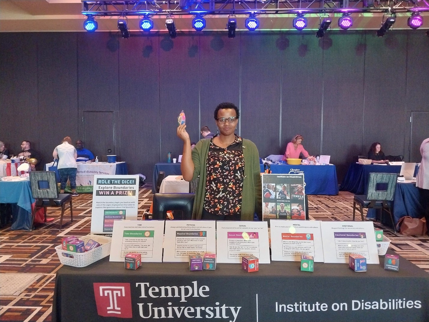 An African woman with short black hair and black glasses stands behind a conference table, that has informational signs on it. She is holding a fidget toy in the hand, lifted up in the air. 