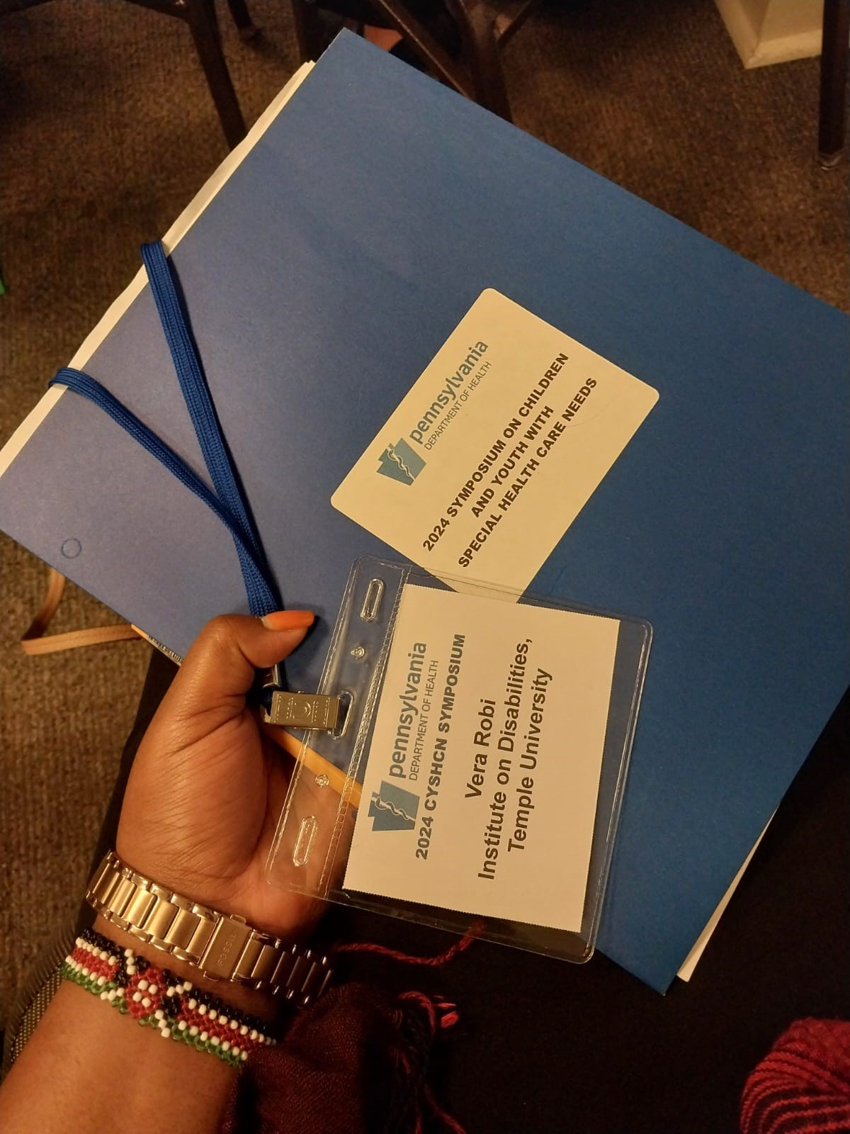 The image includes a woman’s hand, with a watch and bracelet that shows the Tanzanian flag, holding a blue envelope that reads “Pennsylvania Department of Health” 2024 Symposium on Children and Youth With Special Health Care Needs.” On top of the folder is a name card that reads “Vera Robi Institute on Disabilities, Temple University.”