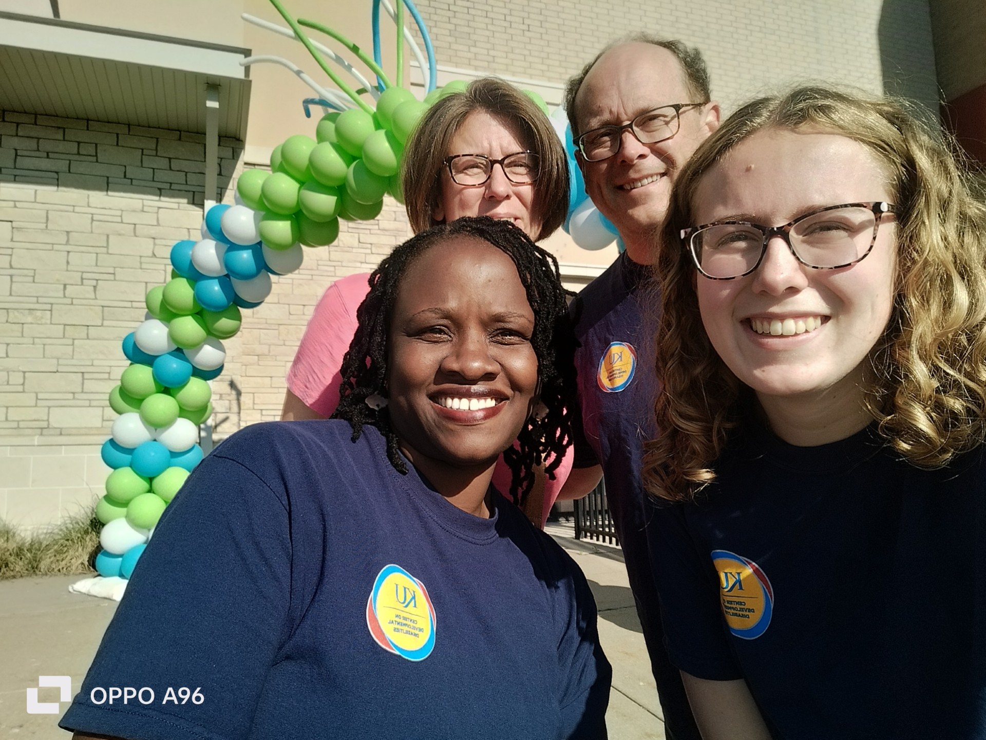 Alt-Text: Four individuals stand outside in front of a balloon arch composed of green, blue, and white balloons. Among them are one African woman, two white women, and one white man, all wearing identical navy blue t-shirts.