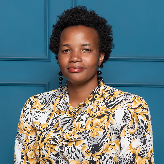 An African woman with black hair is wearing black earrings and a colorful black-brown flower print blouse as she poses for a photo against a blue background.